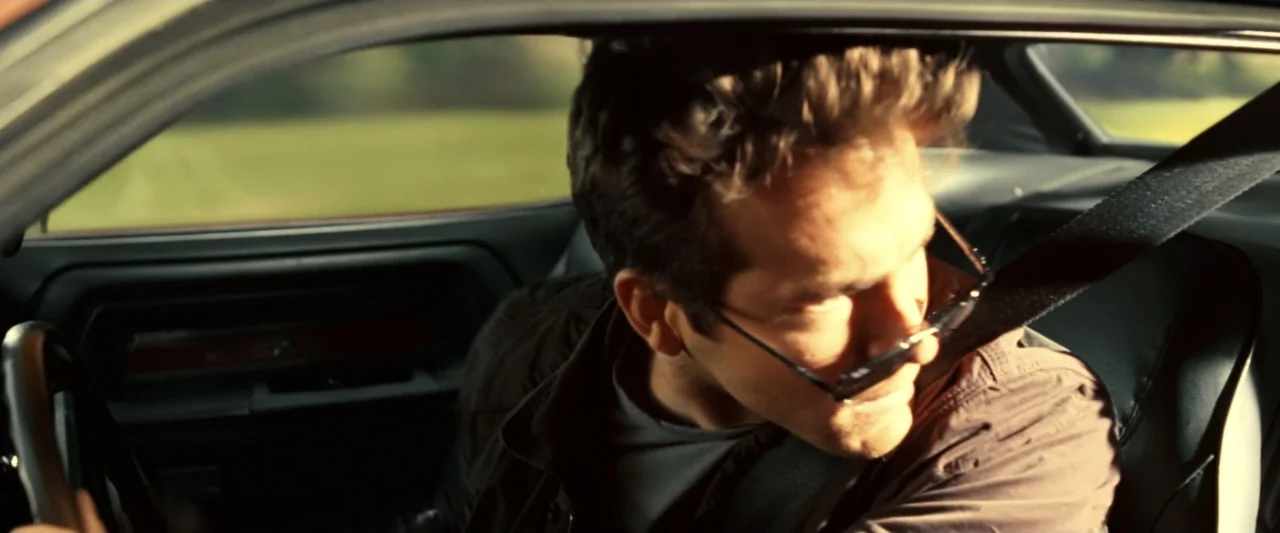 Green Lantern (2011) movie still: medium — The interior of a moving car shows an open door frame and dark seats. A man in a brown shirt turns his…