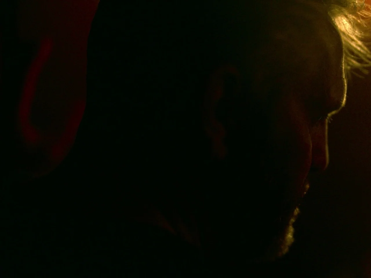 Violet (2014) movie still: closeup — A dark, unidentifiable space features a single source of focused light. A man's face is partially visible in…