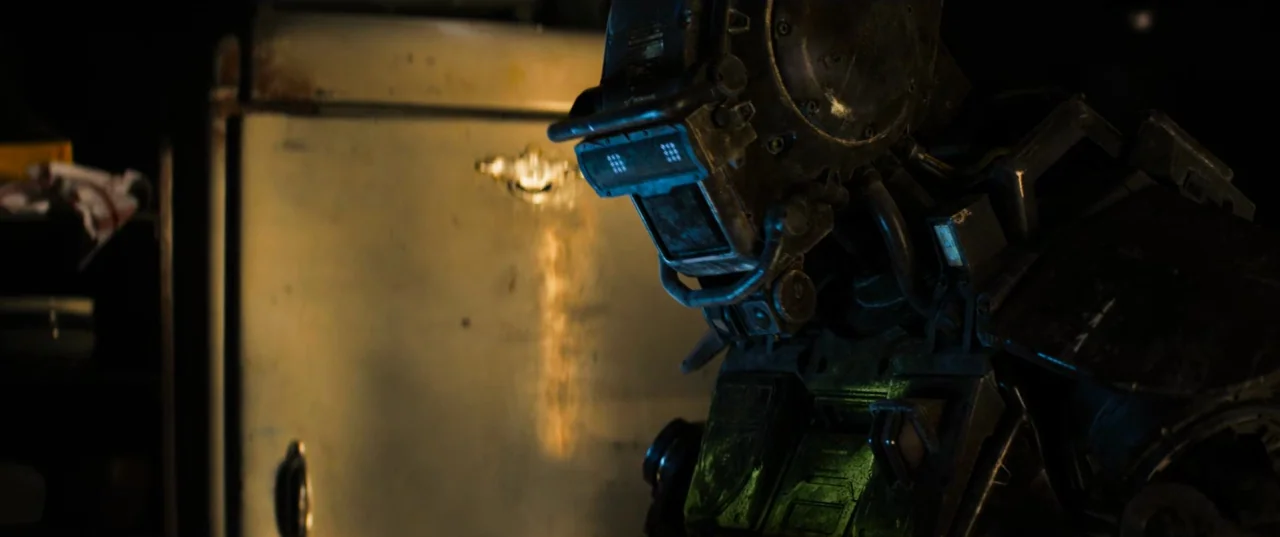 Chappie (2015) movie still: medium — A dark, cluttered industrial space contains a weathered metallic wall. A large, worn-looking robotic figure…