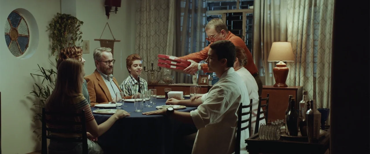 The Boy Who Killed My Parents (2021) movie still: medium — A 1970s dining room is dimly lit by a single table lamp. A group of people sits around a…