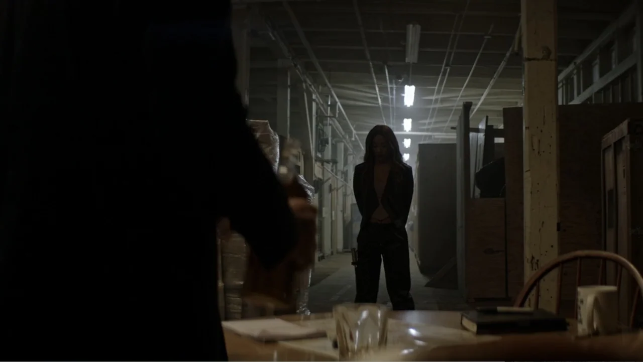 The Collective (2023) movie still: medium — An expansive dim warehouse features wooden partitions and concrete floors stretching into the shadows. A…