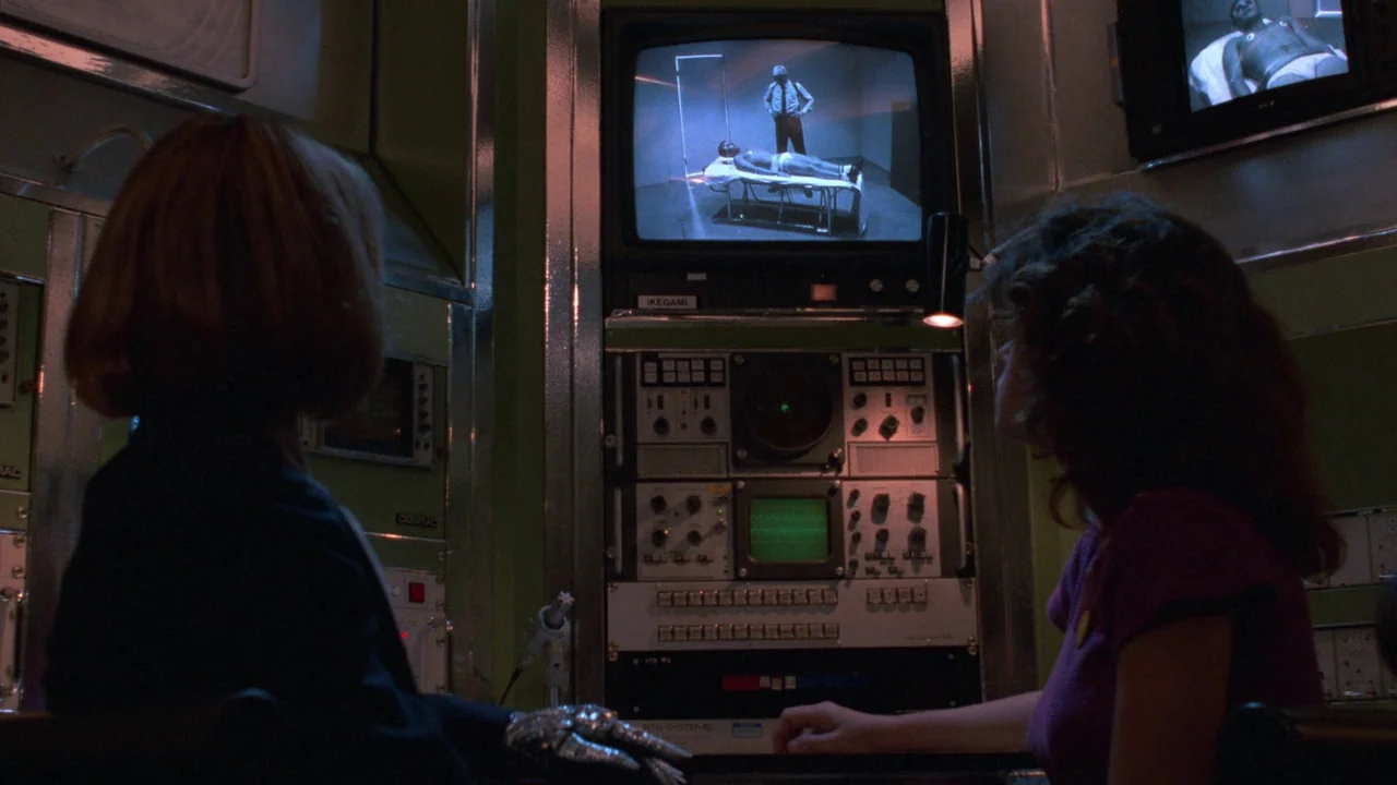 Repo Man (1984) movie still: medium — A dimly lit technical control room features metal walls and stacked video surveillance equipment. Two women…