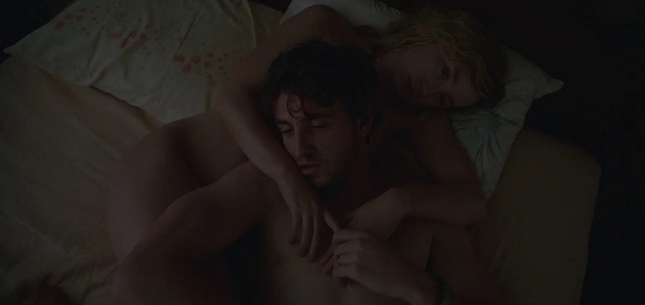 Foe (2023) movie still: medium — A man and woman lie together in a darkened bed with light-colored sheets. The woman is positioned behind the man,…