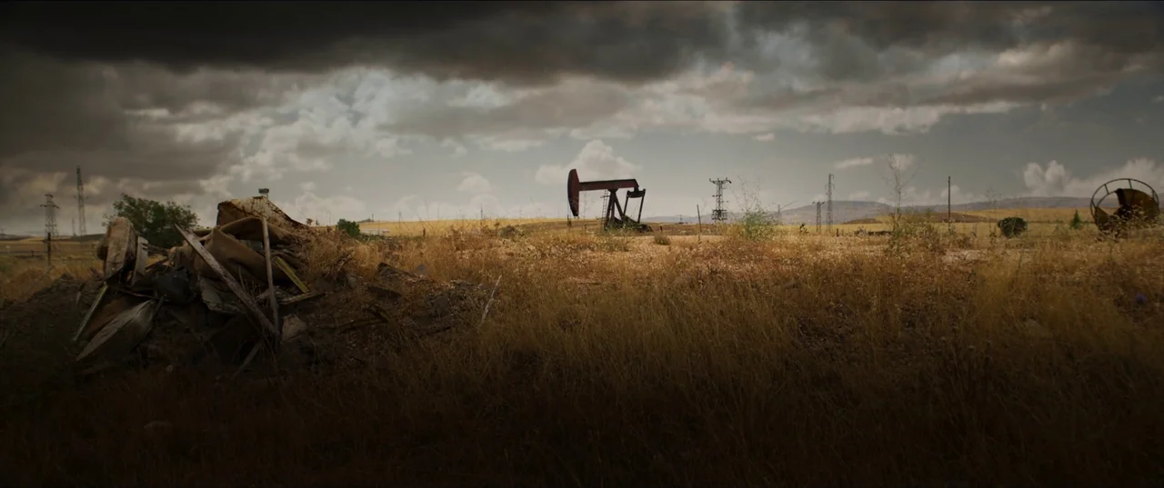 About Dry Grasses (2023) movie still: establishing — A desolate, dry field contains a rusted oil pumpjack and a pile of debris. Overcast, dark…