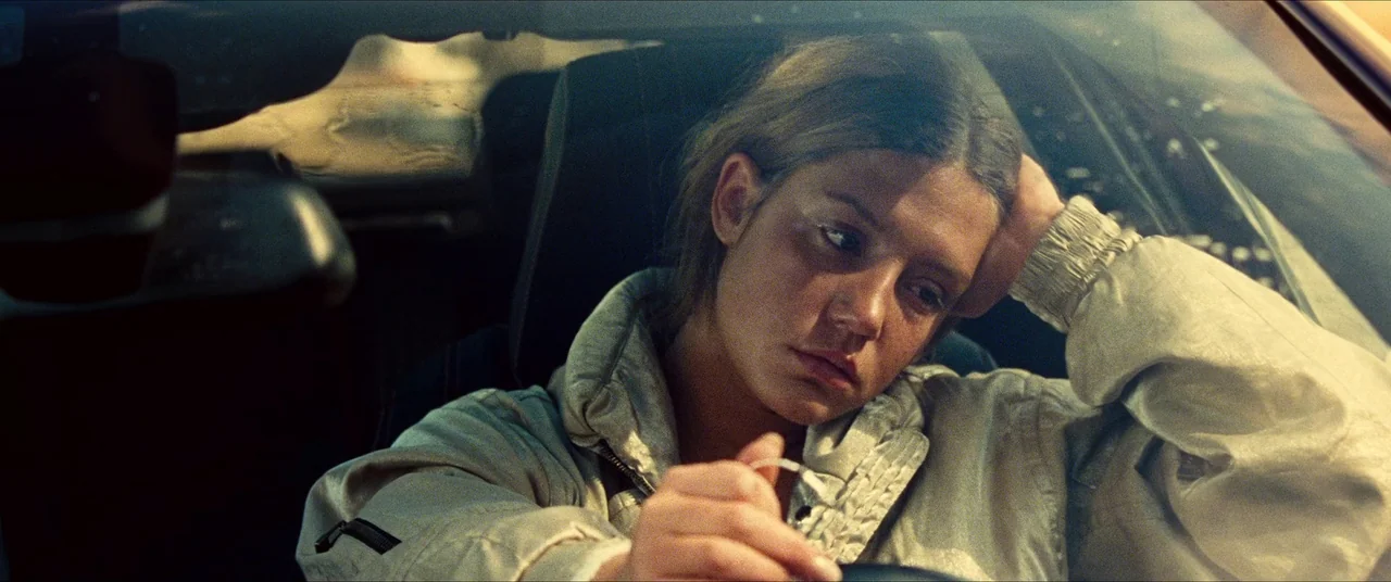 The Five Devils (2022) movie still: medium — A woman sits in the driver seat of a vehicle during daylight hours. She rests her head on her hand…