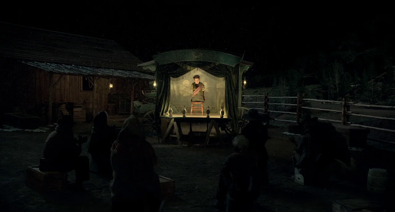 The Ballad of Buster Scruggs (2018) movie still: wide — A night encampment features a wooden stage built onto a wagon with curtains pulled back. A…