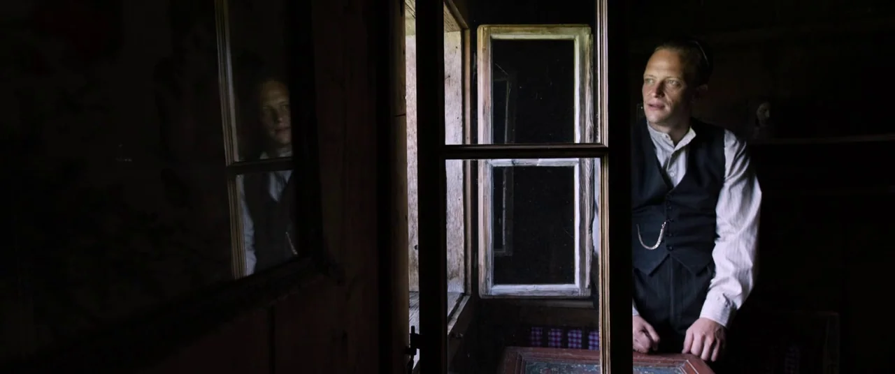 A Hidden Life (2019) movie still: medium — A dimly lit room features rustic wooden paneling and a small rectangular window. A man in a vest stands…
