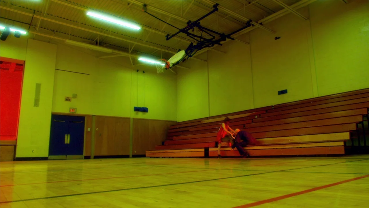 The ABCs of Death (2012) movie still: wide — A high-school basketball court with wooden bleachers and exposed metal ceiling supports. Two people sit…