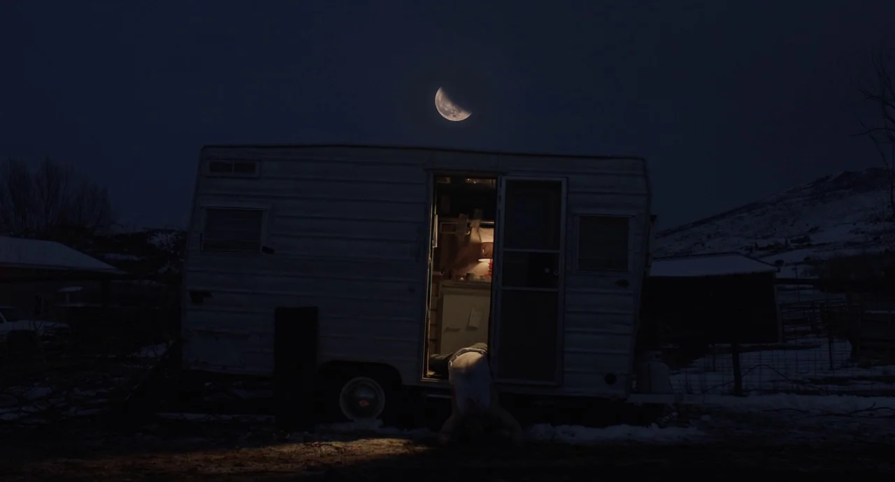 The Wolf of Snow Hollow (2020) movie still: wide — A silver travel trailer sits in a snowy, desolate lot under a crescent moon. A person crawls out…