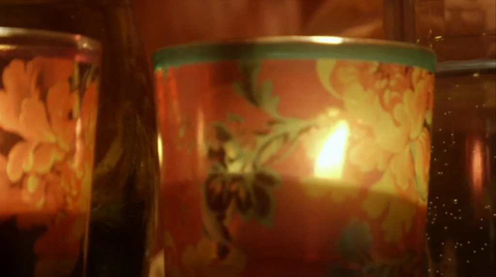 Humpday (2009) movie still: closeup — A close view of a lit candle inside an orange floral patterned glass holder. The glass glows brightly as the…