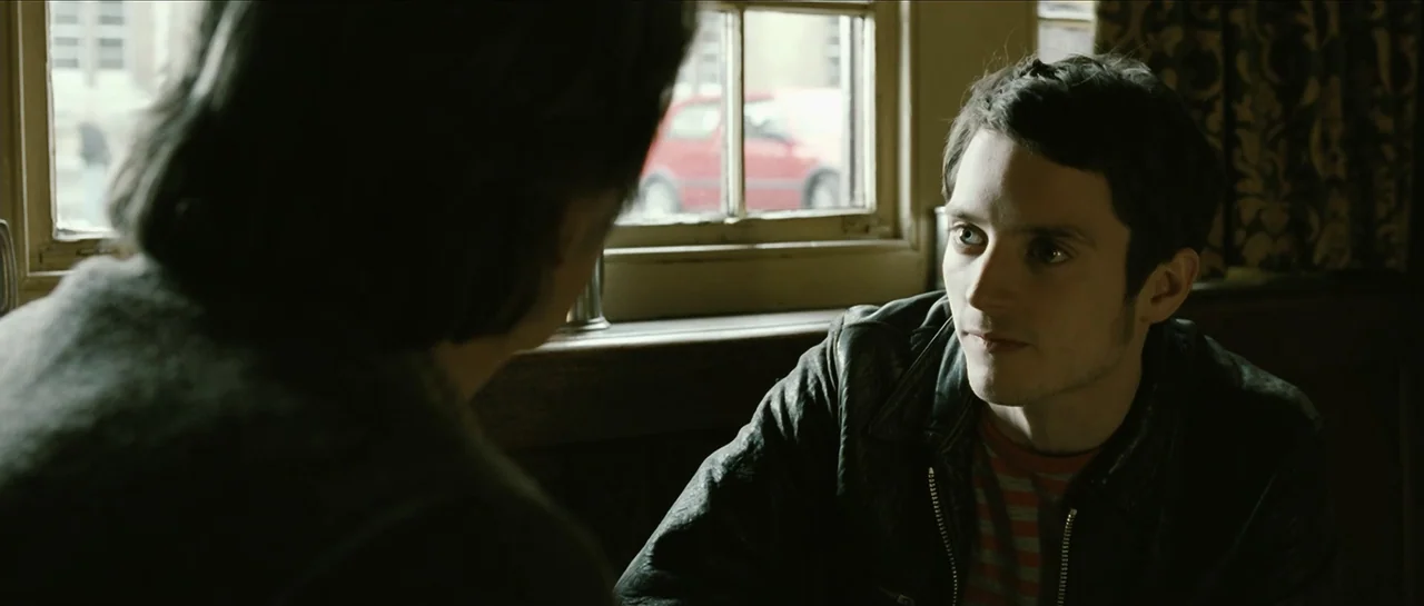 The Oxford Murders (2008) movie still: medium — A dimly lit pub interior features dark wooden paneling and a small window backdrop. A man wearing a…