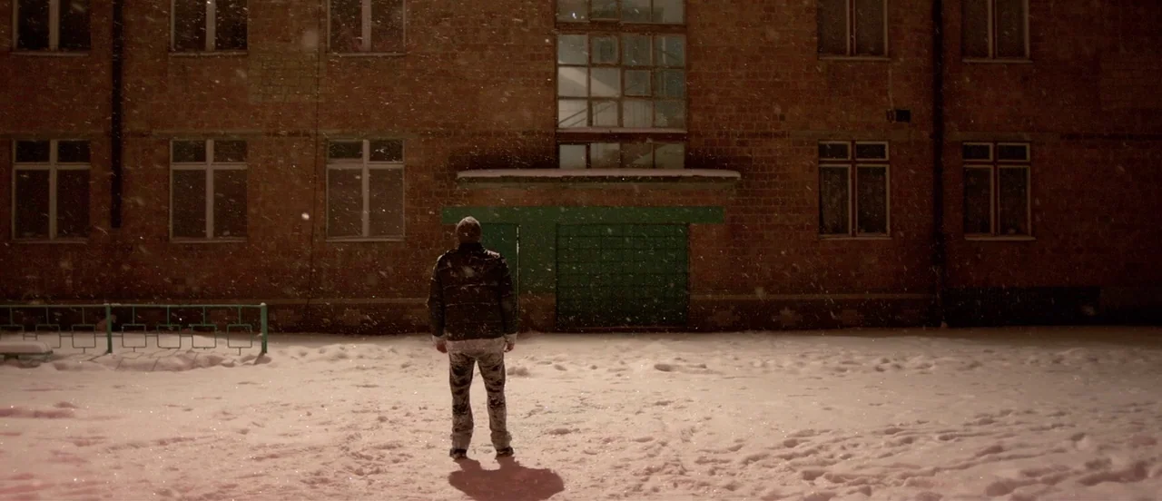 The Tribe (2014) movie still: fullbody — A brick apartment building at night with a snow-covered yard in the foreground. A lone person stands in the…