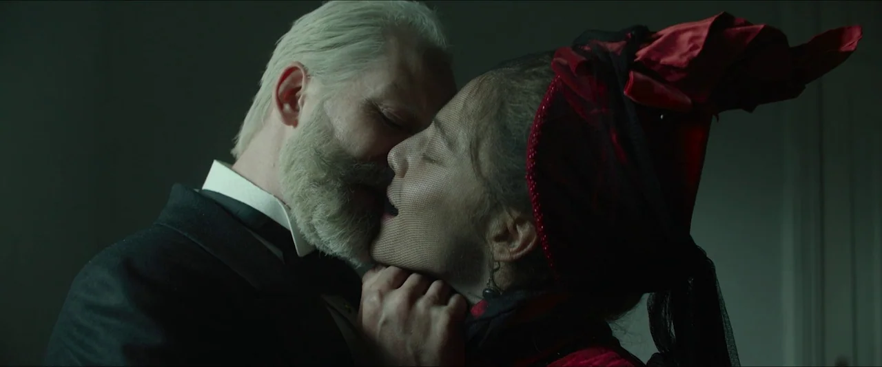 Tchaikovsky’s Wife (2022) movie still: closeup — A dimly lit interior space with a neutral-toned wall background. A man with a white beard kisses a…