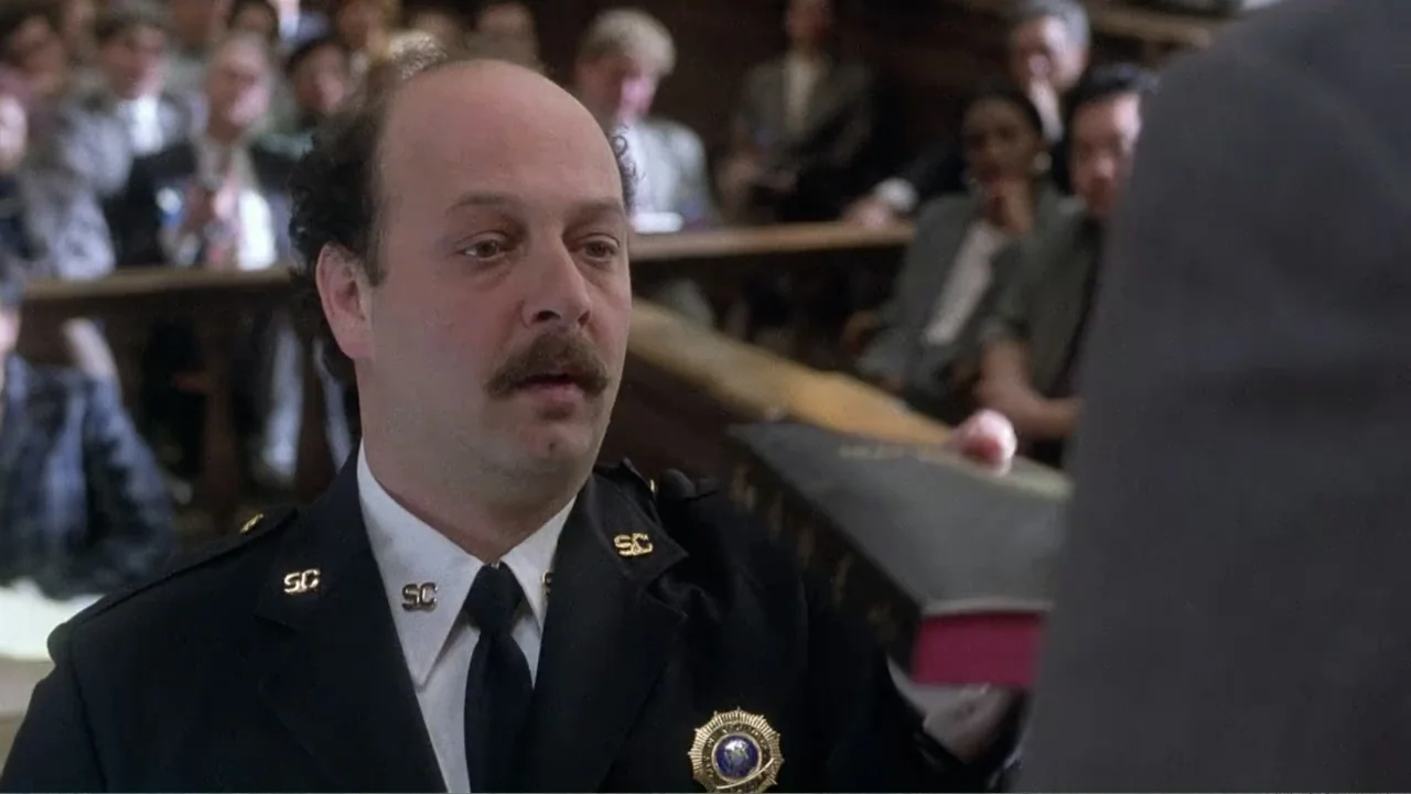 Mistrial (1996) movie still: medium — A wood-paneled courtroom filled with blurred people seated in the background pews. A police officer in a dress…