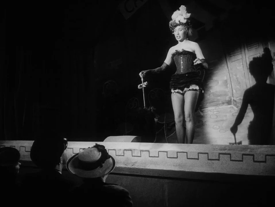 A Ship Bound for India (1947) movie still: fullbody — A dark cabaret stage features a performer in a corset and feathered headpiece. The foreground…