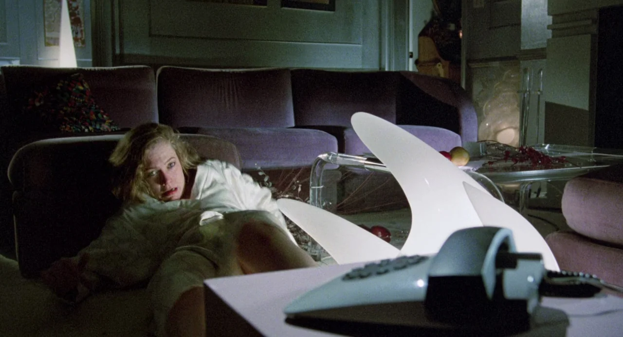 Crimes of Passion (1984) movie still: medium — A dark living room features a purple velvet sofa and a low, white sculptural lamp. A blonde woman in…