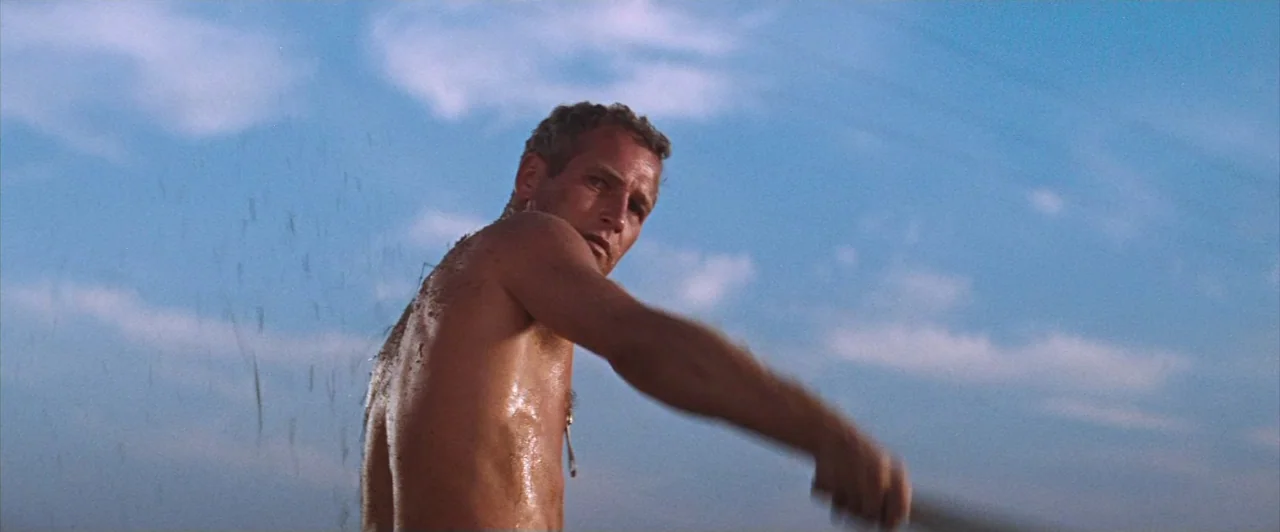 Cool Hand Luke (1967) movie still: medium — An outdoor expanse with a clear blue sky and thin, wispy clouds. A shirtless man turns his torso while…
