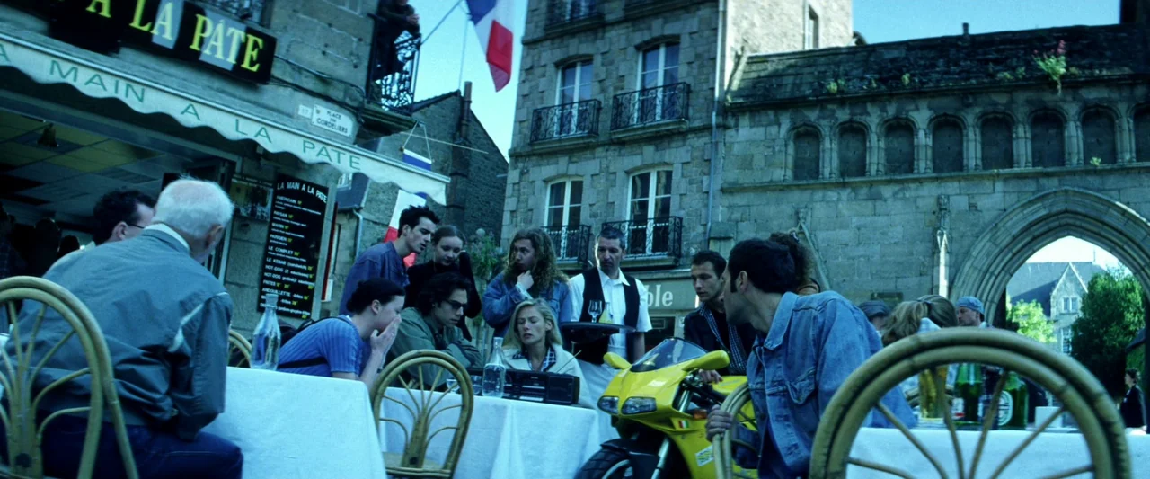 Armageddon (1998) movie still: wide — A European square with stone buildings and a restaurant seating area outdoors. A group of people occupy tables…