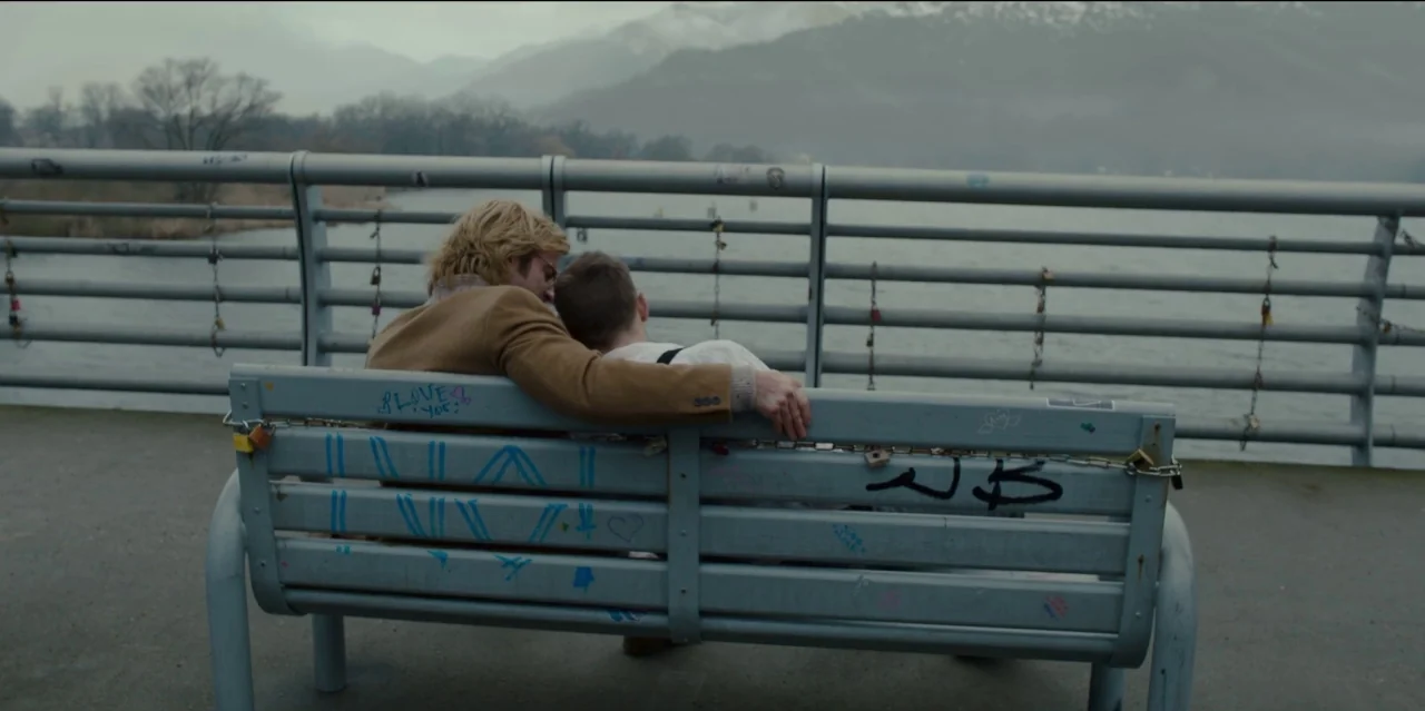 Mute (2018) movie still: wide — A metal bridge overlooks a grey lake with hazy, distant mountains in the background. Two young men sit together on a…