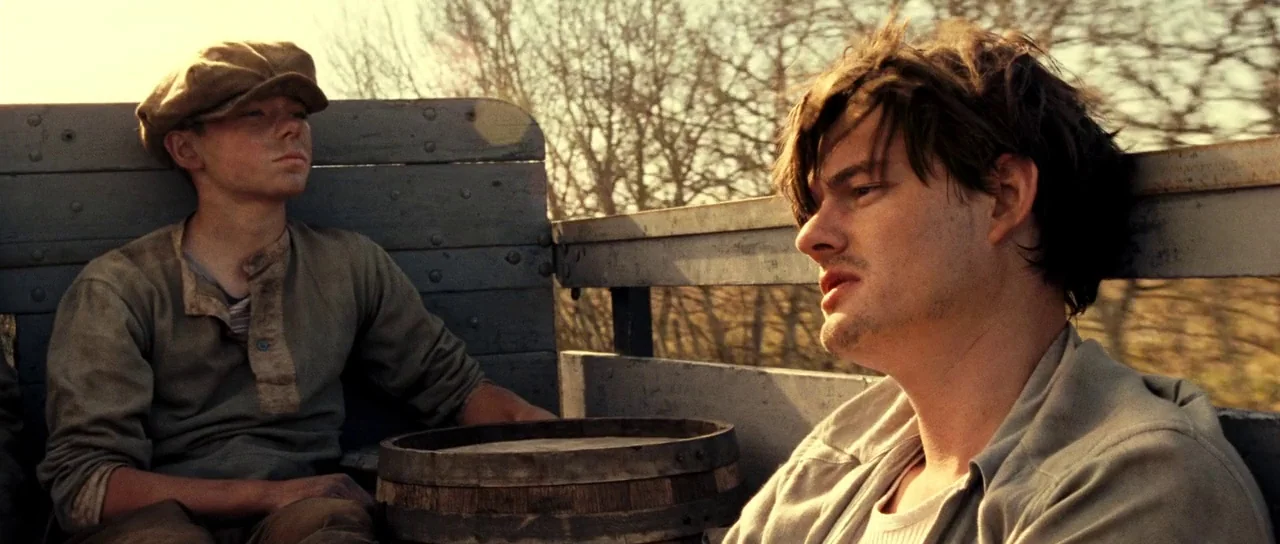 On The Road (2012) movie still: medium — Two men sit side-by-side in the wooden bed of a moving truck. The background consists of blurry trees…