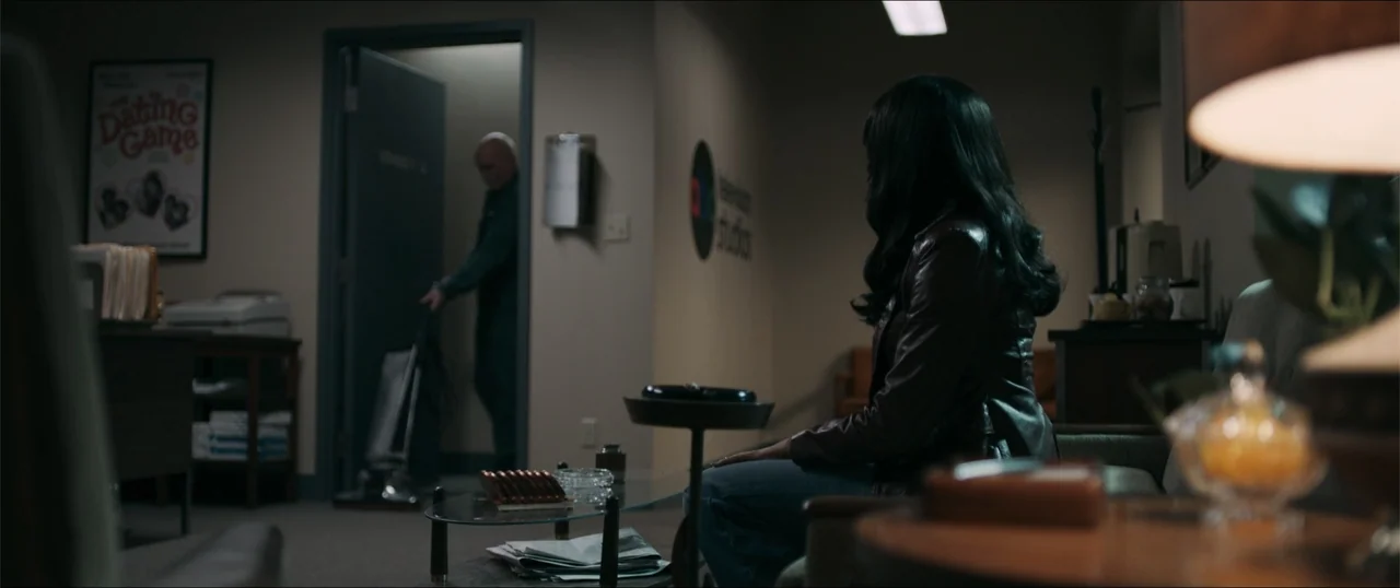 Woman of the Hour (2024) movie still: medium — A dimly lit waiting area features a poster on the wall and a glass coffee table. A woman sits on a…