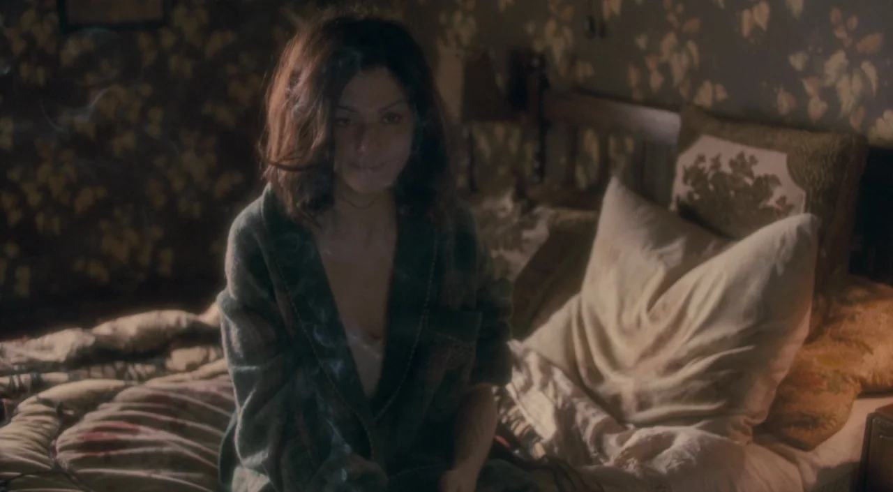 The Deep Blue Sea (2011) movie still: medium — A dimly lit room with vintage patterned wallpaper features a woman sitting on bedding. The woman…