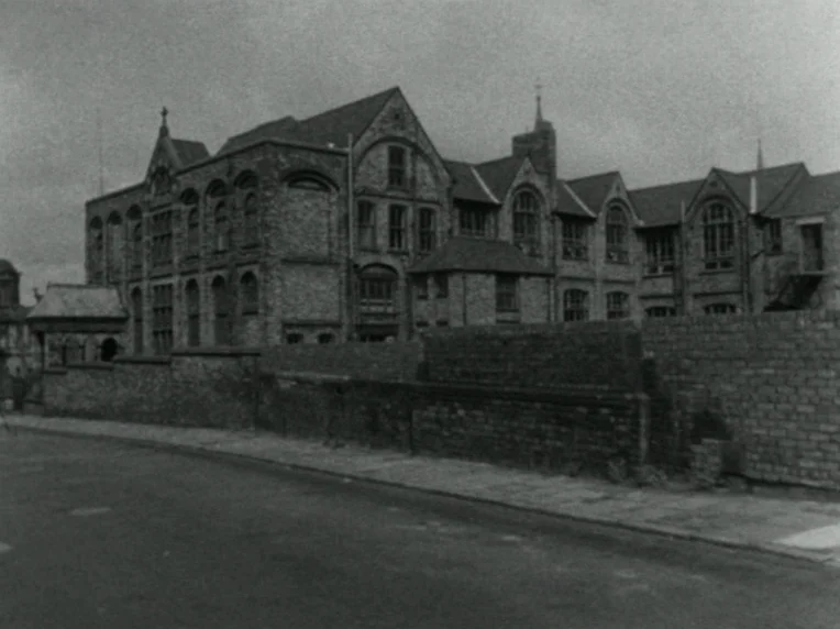 Children (1976) movie still: establishing — A multi-story brick institutional building with numerous arched windows stands behind a low wall. The…