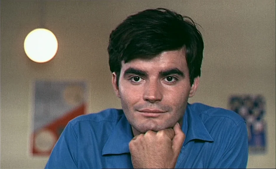 Le Bonheur (Happiness) (1965) movie still: closeup — A man stands in a room with beige walls and blurred posters on either side. The man wears a…