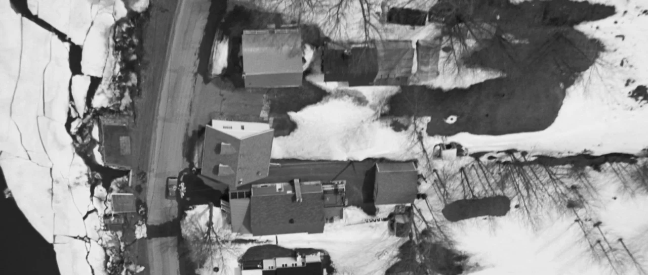 Polytechnique (2009) movie still: establishing — An aerial view shows houses and roads surrounded by patches of melting snow. Bare trees cast thin…