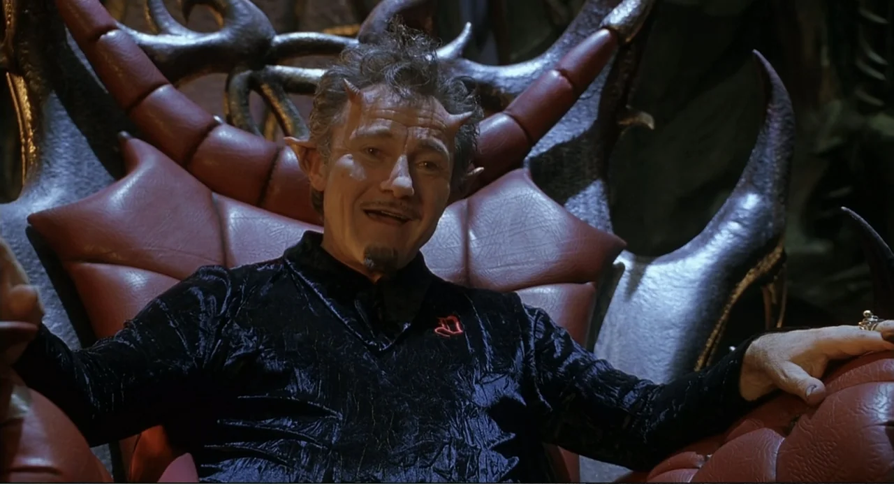 Little Nicky (2000) movie still: medium — A dark space features a man sitting on a large, red-cushioned throne. The man displays demonic facial…