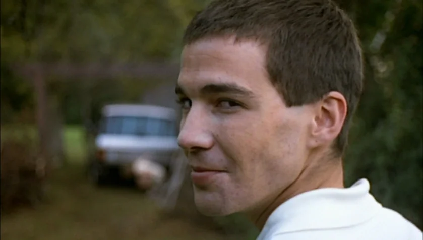 Funny Games (1997) movie still: closeup — An outdoor rural setting features a grass lawn with a parked vehicle in the background. A young man in a…