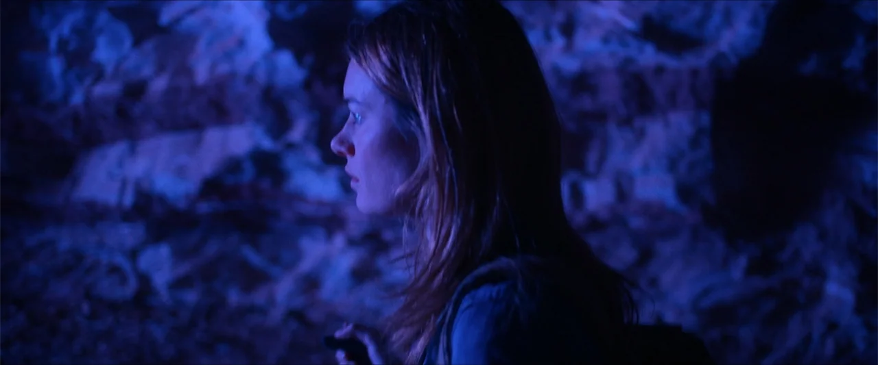 An Endless Wedding (2025) movie still: closeup — A dark rocky cave interior with irregular walls illuminated by dim blue light. A young woman stands…
