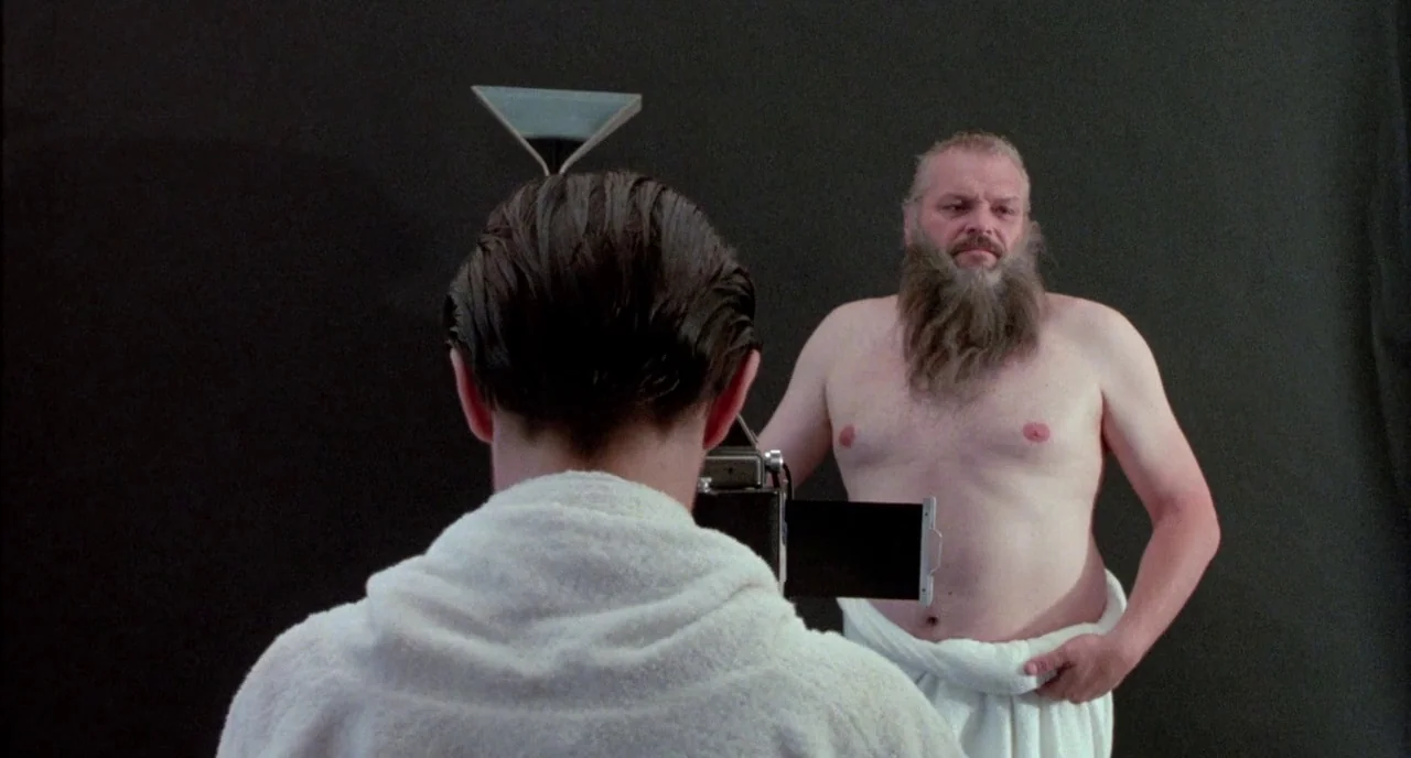 The Belly of an Architect (1987) movie still: medium — A studio space with a plain black background and minimal furniture. An older man wearing only…