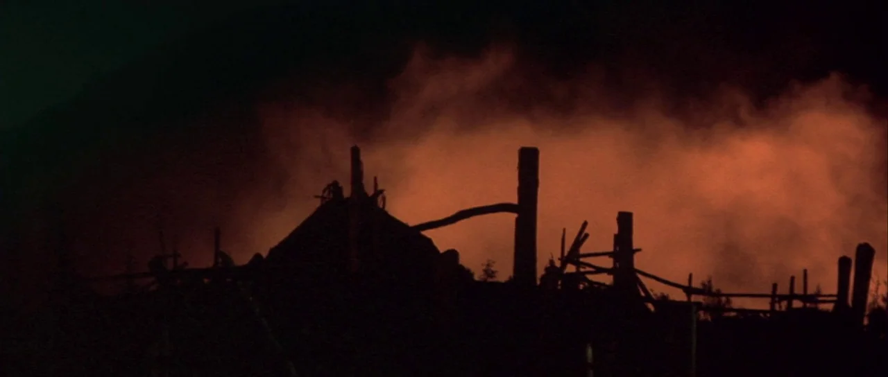 The 13th Warrior (1999) movie still: establishing — A wooden structure with a fence is silhouetted against a wall of fire. Dark smoke and embers…