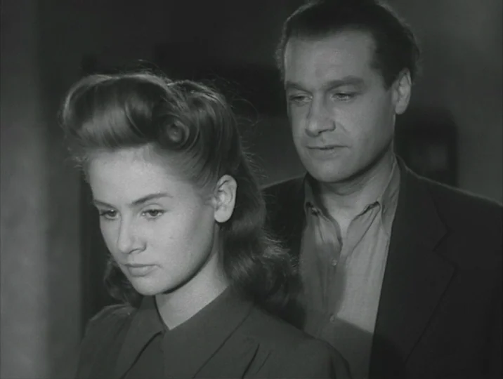 Crisis (1946) movie still: medium — A dark, minimally lit interior space serves as the backdrop for the two figures. A young woman and a man in a…