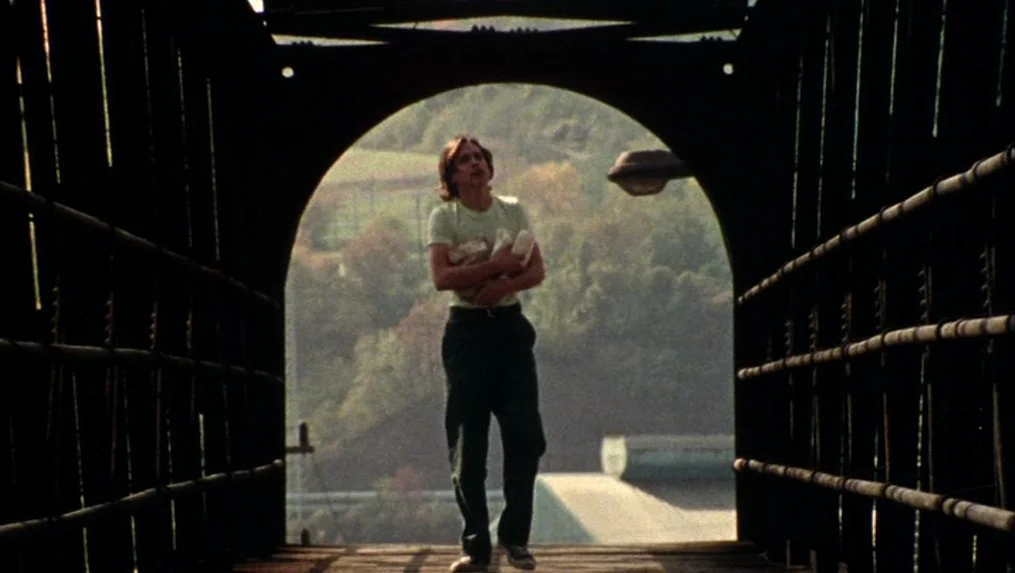 Martin (1977) movie still: fullbody — A metal bridge with an arched opening frames a person in the center. The man walks forward holding a bundle…