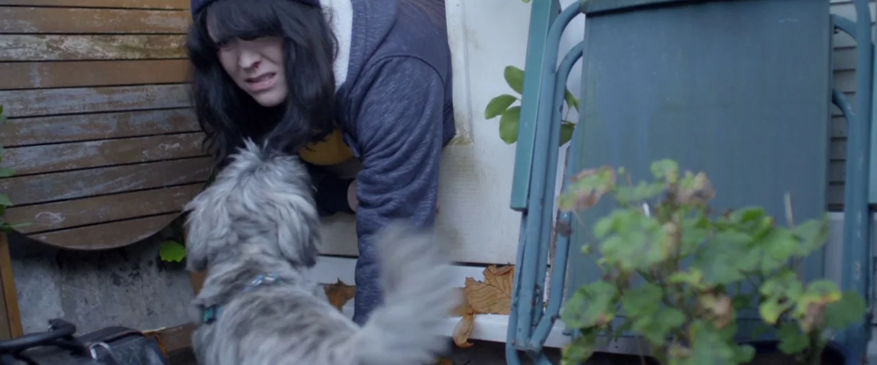Prevenge (2016) movie still: medium — A residential porch area with wood siding and a folded green chair visible. A woman looks down at a small…