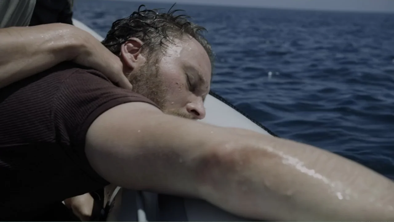 Capsized: Blood in the Water (2019) movie still: closeup — A man lies unconscious across the edge of an inflatable boat on open water. A second…