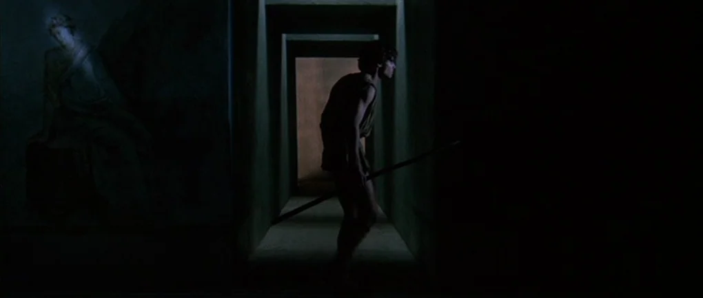 Fellini – Satyricon (1969) movie still: medium — A narrow dark stone corridor features a faded classical fresco of a seated figure. A young man in a…