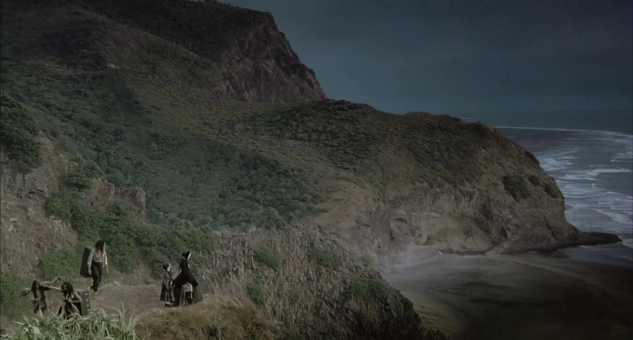 The Piano (1993) movie still: wide — A rugged cliffside overlooks a dark ocean beach under a heavy, overcast sky. Four people carry heavy wooden…