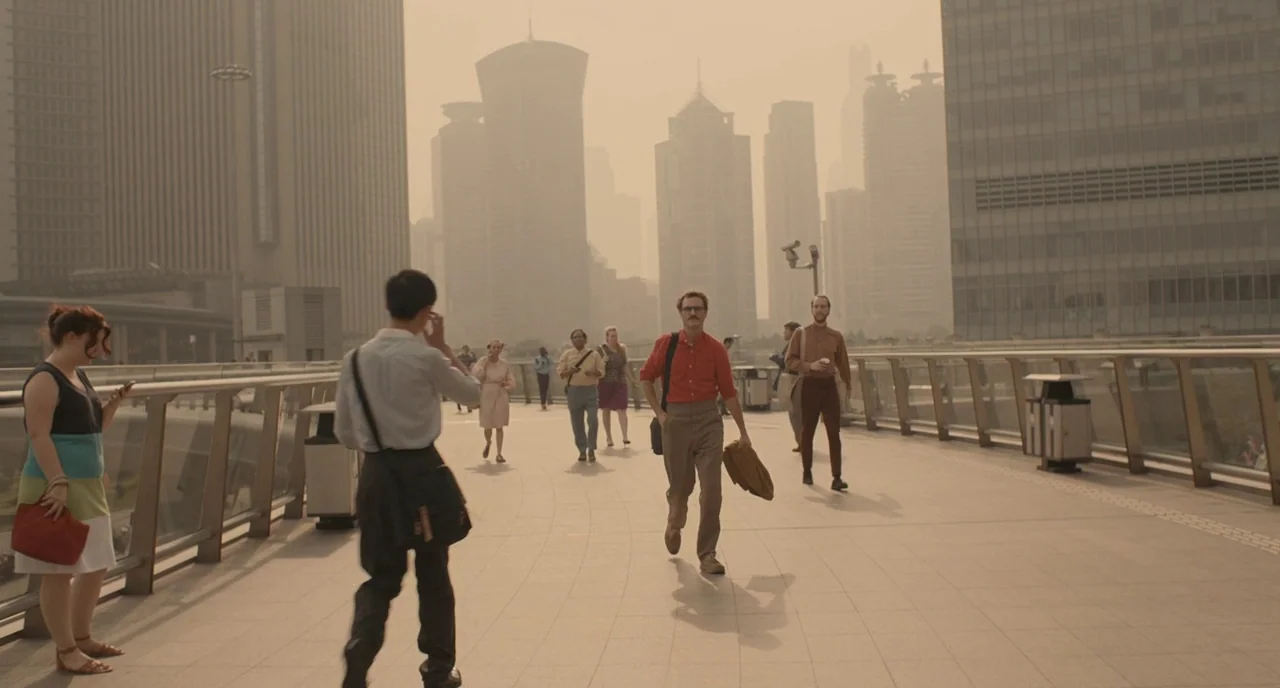 Her (2013) movie still: wide — A pedestrian walkway connects tall glass skyscrapers under a hazy, golden, smoggy sky. Commuters walk in different…