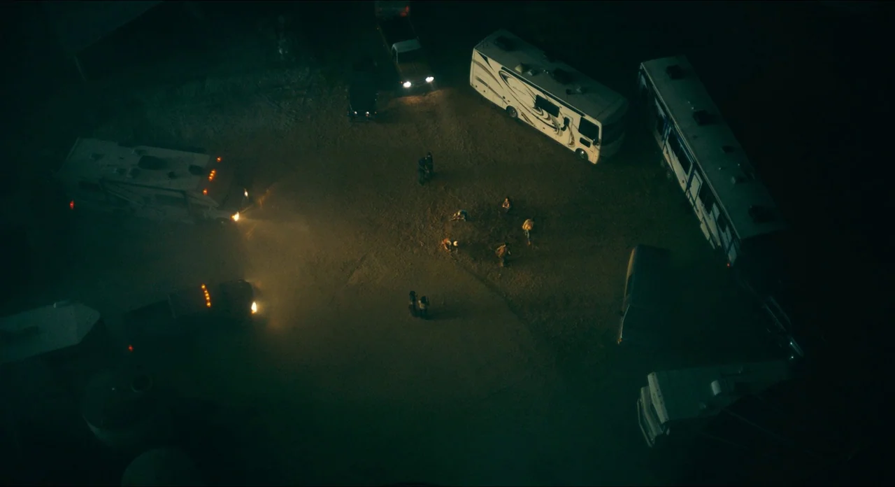 Doctor Sleep movie still: wide — A dark, unpaved dirt lot contains several parked recreational vehicles and trucks at night. A small group of people…