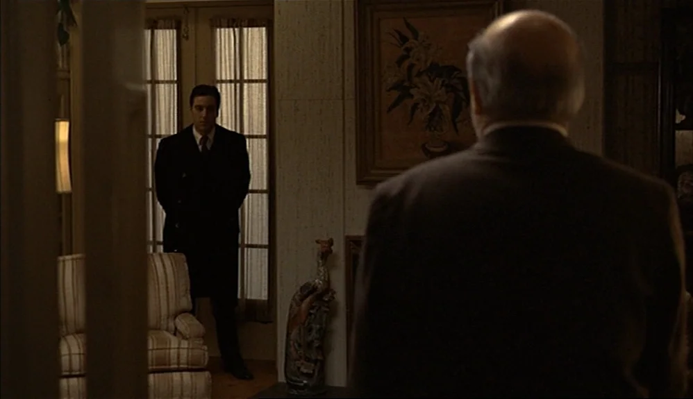 The Godfather Part II (1974) movie still: medium — A dimly lit room with wood-paneled walls features a floral painting and patterned furniture. One…