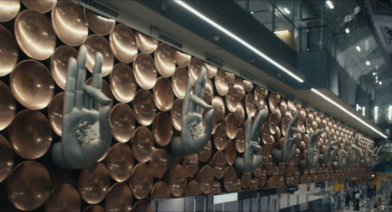 Origin (2023) movie still: wide — A wall in an airport terminal is decorated with repetitive copper circular plates and sculpted hands in mudras.…