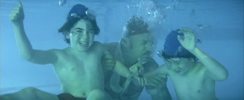 The Royal Tenenbaums (2001) movie still: medium — An underwater perspective of a swimming pool with murky, blue-tinted water. A man holds two young…