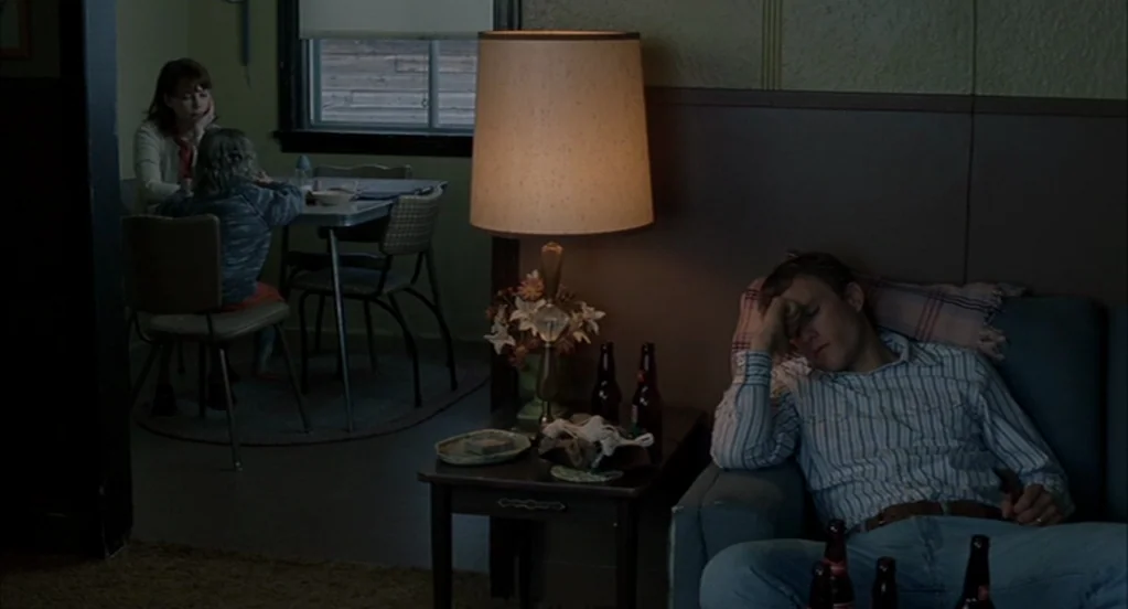 Brokeback Mountain (2005) movie still: wide — A dimly lit room features a man resting on a blue couch beside a lamp. In the background, a woman and…