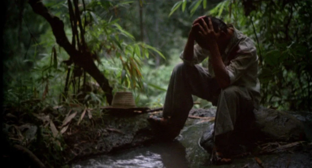 The Killing Fields (1984) movie still: fullbody — A dense jungle setting features lush green foliage and a small rocky creek. A man sits by the…