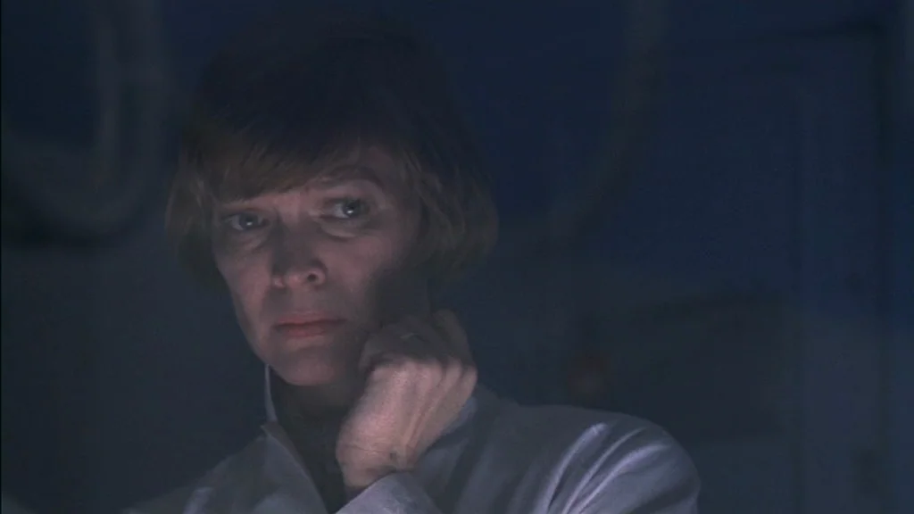 The Exorcist (1973) movie still: closeup — A dark, dimly lit interior space features blue atmospheric lighting and industrial textures. A woman with…