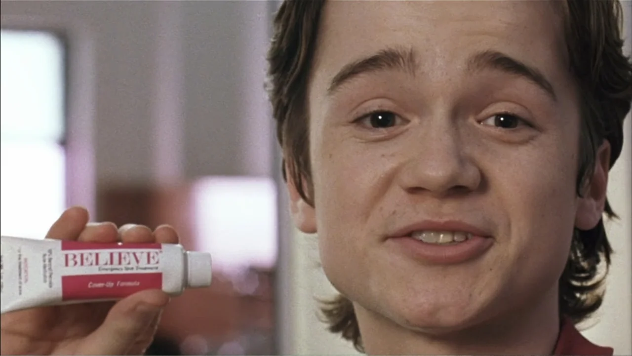 A Cinderella Story (2004) movie still: closeup — A brightly lit interior space with an out-of-focus background. A teenage boy holds up a tube of…