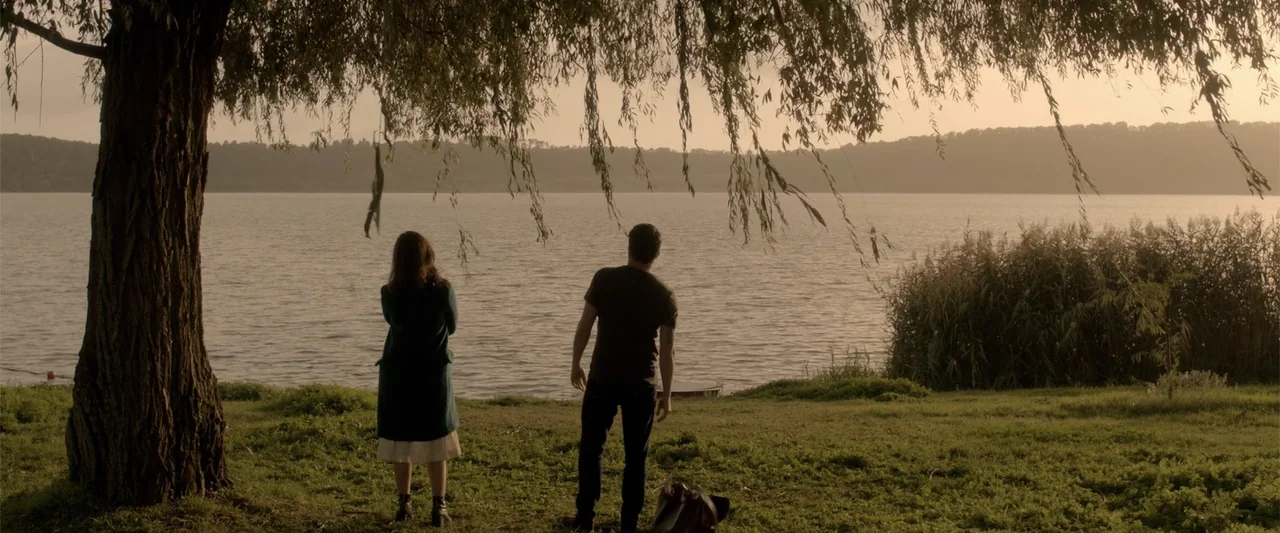 About Us (2017) movie still: fullbody — A lakeside grassy bank with a large willow tree in the foreground. A man and woman stand side by side…